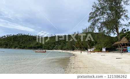 Tropical island coastline, white sand, clear water, tourists walking on the beach, beach bar and boat on the water, mountains with palm trees in the background. 121386262