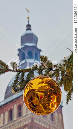 Glowing golden bauble reflecting holiday festivities, framed by snow-covered evergreen branch and historic clock tower in background Glowing golden bauble reflecting holiday festivities, framed by snow-covered evergreen branch and historic clock tower in background 121386434