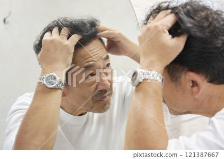 一個 50 多歲的男人看著鏡子，擔心自己的白髮。 121387667