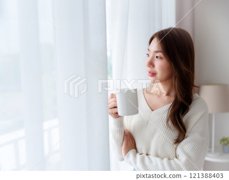 Young asian woman standing beside window and holding mug in bedroom at home 121388403
