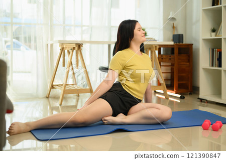 Attractive young woman practicing pigeon pose on blue exercise mats in living room 121390847