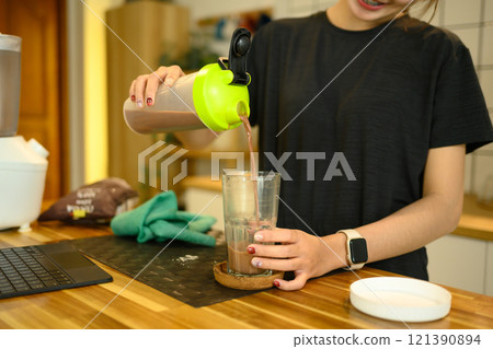 Smiling woman in sportswear pouring a freshly made chocolate protein shake into a glass 121390894