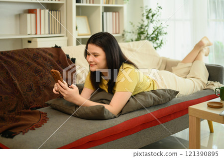 Young woman in casual clothes laying on a couch and using smartphone 121390895