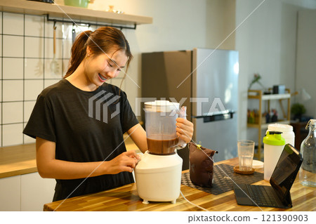 Cheerful young woman blending chocolate protein shake or health drink with blender at kitchen counter 121390903