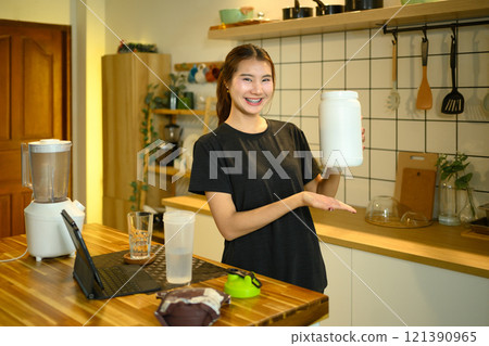 Smiling asian woman holding protein powder container standing in warm kitchen 121390965