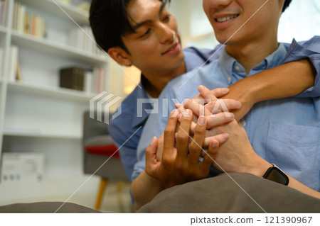 Loving gay couple holding hands and smiling while sharing a warm embrace on sofa 121390967