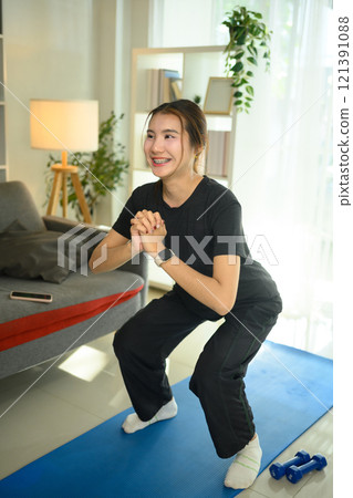 Beautiful young woman doing squat exercise on mat in her living room. Active and healthy lifestyle Beautiful young woman doing squat exercise on mat in her living room. Active and healthy lifestyle 121391088