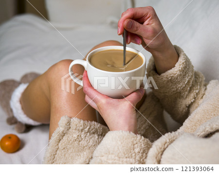 Woman in a bathrobe is sitting on a white bed, stirring a cup of coffee with a spoon. There are tangerines scattered on the bed around her 121393064