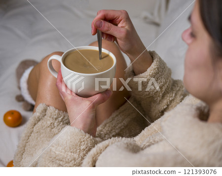 Young woman enjoys a cozy morning at home, wearing a beige bathrobe and stirring her coffee with a metal spoon, embracing relaxation during breakfast Young woman enjoys a cozy morning at home, wearing a beige bathrobe and stirring her coffee with a metal spoon, embracing relaxation during breakfast 121393076
