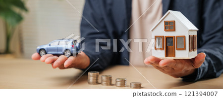 A person holds a car and house model above coin stacks, symbolizing real estate, auto loans, savings, and financial goals 121394007