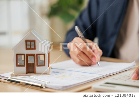 A man writes a signature on house insurance documents, using a calculator for tax and property details. Counting payments and rent by model house, he finalizes the price A man writes a signature on house insurance documents, using a calculator for tax and property details. Counting payments and rent by model house, he finalizes the price 121394012