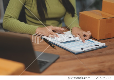 Startup SME small business entrepreneur of freelance Asian woman wearing apron using laptop and box to receive and review orders online to prepare to pack sell to customers, online sme business ideas. 121395228