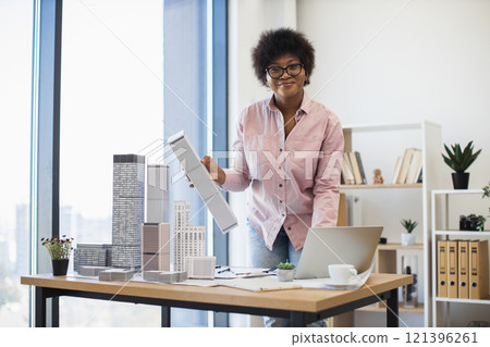 African American female architect in casual attire working on city models. Bright workspace with large windows enhances creativity and focus. African American female architect in casual attire working on city models. Bright workspace with large windows enhances creativity and focus. 121396261