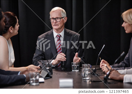 Senior politician in formal wear chairing committee answering questions during press conference while sitting at table with other administration officials Senior politician in formal wear chairing committee answering questions during press conference while sitting at table with other administration officials 121397465