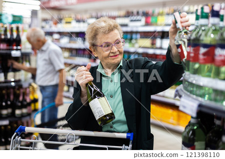 Mature woman with glasses chooses bottle of wine in alcohol section of supermarket 121398110