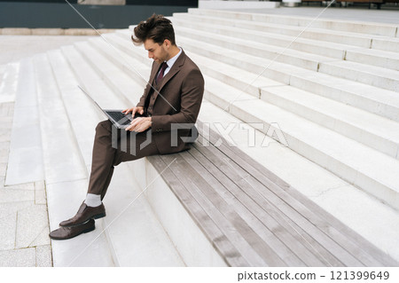 Wide shot of focused businessman working remotely while sitting on steps in urban setting, typing on laptop computer, focused looking to screen. Concept of business communication and technology. Wide shot of focused businessman working remotely while sitting on steps in urban setting, typing on laptop computer, focused looking to screen. Concept of business communication and technology. 121399649