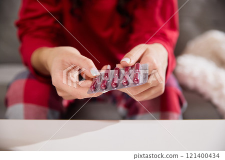 Cropped photo of young female hands holding blister pack of red capsules, carefully examining them over white table. Cropped photo of young female hands holding blister pack of red capsules, carefully examining them over white table. 121400434