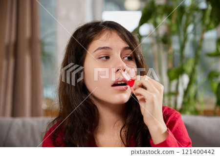 Close-up of young woman holding red lozenge near her mouth, about to take medication, with focused expression. Close-up of young woman holding red lozenge near her mouth, about to take medication, with focused expression. 121400442