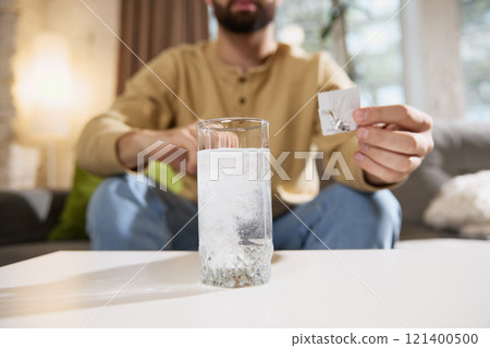 Cropped photo of young man feels bad, sick and unpack effervescent tablet and put it to glass of water sitting on sofa at home. Cropped photo of young man feels bad, sick and unpack effervescent tablet and put it to glass of water sitting on sofa at home. 121400500
