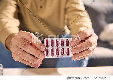 Young sick, feels badly man in casual beige shirt holds blister pack of red capsules, sitting in bright, cozy living room. Young sick, feels badly man in casual beige shirt holds blister pack of red capsules, sitting in bright, cozy living room. 121400502