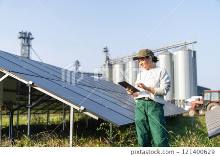 Female farmer with digital tablet on a modern farm using solar panels. Agricultural silos in the background Female farmer with digital tablet on a modern farm using solar panels. Agricultural silos in the background 121400629