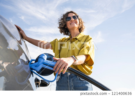 Woman with fueling hydrogen car. Refueling car with hydrogen fuel. Wind turbines in the background Woman with fueling hydrogen car. Refueling car with hydrogen fuel. Wind turbines in the background 121400636