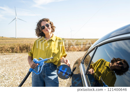 Woman holds a hydrogen fueling nozzle. Refueling car with hydrogen fuel. Wind turbines in the background. Woman holds a hydrogen fueling nozzle. Refueling car with hydrogen fuel. Wind turbines in the background. 121400646