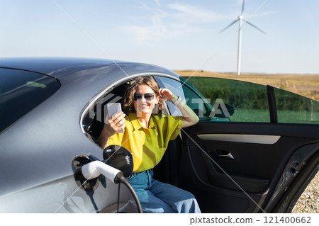 Woman with curve hairs wearing yellow shirt and sunglasses sits into charging electric car and holds smartphone. Wind turbines in the background Woman with curve hairs wearing yellow shirt and sunglasses sits into charging electric car and holds smartphone. Wind turbines in the background 121400662