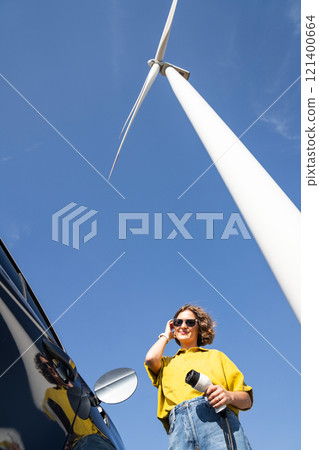 Woman with curve hairs wearing yellow shirt and sunglasses stands next to charging electric car. Wind turbine in the background. Woman with curve hairs wearing yellow shirt and sunglasses stands next to charging electric car. Wind turbine in the background. 121400664