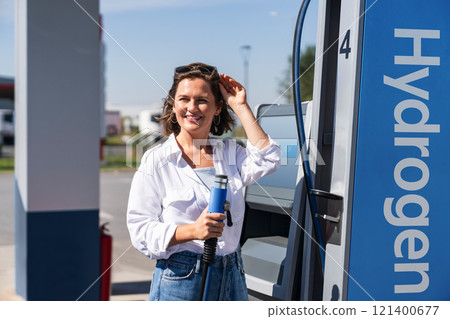 Woman holds a hydrogen fueling nozzle on a hydrogen filling station. Refueling car with hydrogen fuel. 121400677