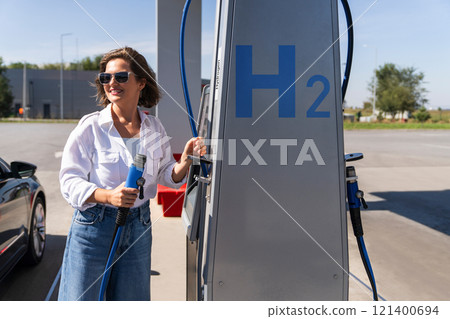 Woman holds a hydrogen fueling nozzle on a hydrogen filling station. Refueling car with hydrogen fuel Woman holds a hydrogen fueling nozzle on a hydrogen filling station. Refueling car with hydrogen fuel 121400694