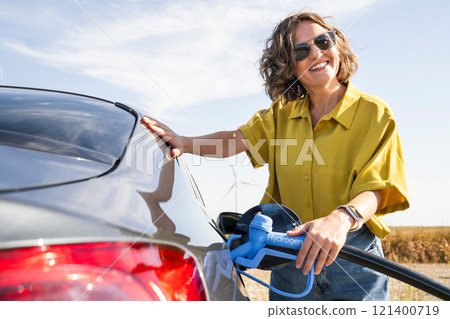Woman with fueling hydrogen car. Refueling car with hydrogen fuel. Wind turbines in the background Woman with fueling hydrogen car. Refueling car with hydrogen fuel. Wind turbines in the background 121400719