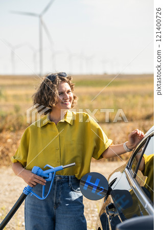 Woman holds a hydrogen fueling nozzle. Refueling car with hydrogen fuel. Wind turbines in the background Woman holds a hydrogen fueling nozzle. Refueling car with hydrogen fuel. Wind turbines in the background 121400726