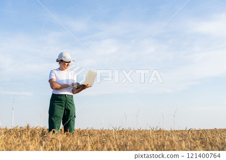 Woman farmer wearing white cap and t-shirt with laptop on agricultural field at sunset. Wind turbines in the background.. Woman farmer wearing white cap and t-shirt with laptop on agricultural field at sunset. Wind turbines in the background.. 121400764