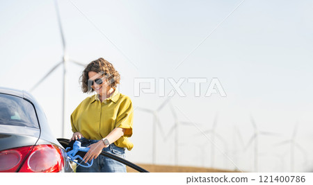 Woman holds a hydrogen fueling nozzle. Refueling car with hydrogen fuel. Wind turbines in the background Woman holds a hydrogen fueling nozzle. Refueling car with hydrogen fuel. Wind turbines in the background 121400786