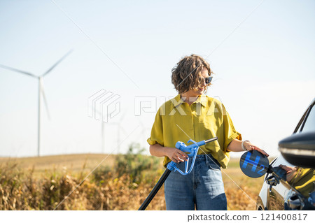 Woman holds a hydrogen fueling nozzle. Refueling car with hydrogen fuel. Wind turbines in the background. 121400817