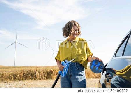 Woman holds a hydrogen fueling nozzle. Refueling car with hydrogen fuel. Wind turbines in the background. Woman holds a hydrogen fueling nozzle. Refueling car with hydrogen fuel. Wind turbines in the background. 121400832