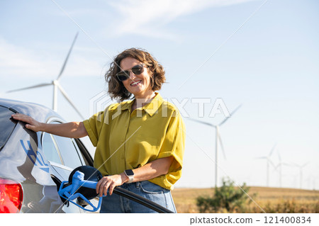 Woman holds a hydrogen fueling nozzle. Refueling car with hydrogen fuel. Wind turbines in the background. Woman holds a hydrogen fueling nozzle. Refueling car with hydrogen fuel. Wind turbines in the background. 121400834