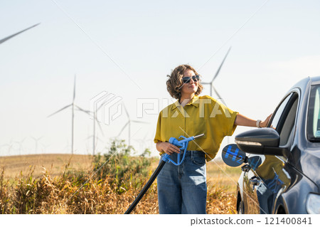 Woman holds a hydrogen fueling nozzle. Refueling car with hydrogen fuel. Wind turbines in the background. Woman holds a hydrogen fueling nozzle. Refueling car with hydrogen fuel. Wind turbines in the background. 121400841