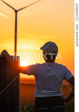 Woman wearing white cap stands next to solar panel at sunset. Wind turbines in the background. Woman wearing white cap stands next to solar panel at sunset. Wind turbines in the background. 121400914