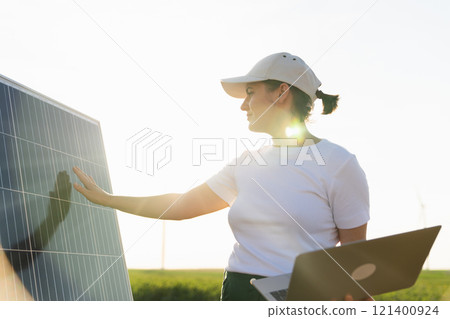 Woman farmer wearing white cap and t-shirt with laptop stands next to solar panel. 121400924