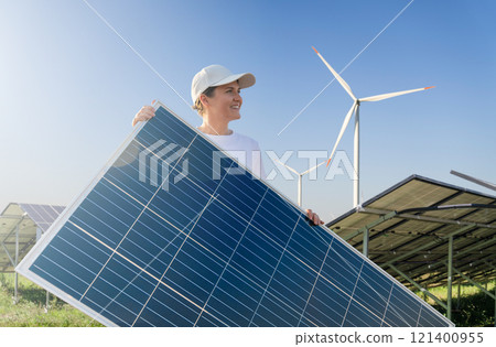 Woman wearing white cap and t-shirt holds solar panel. Wind turbines in the background 121400955