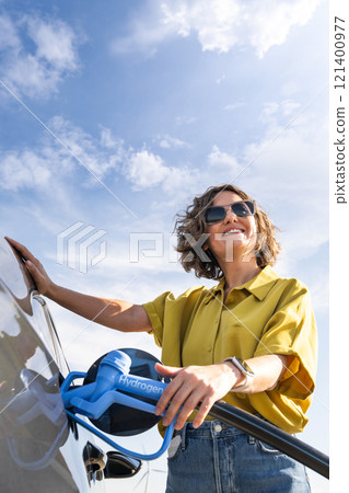 Woman with fueling hydrogen car. Refueling car with hydrogen fuel. Wind turbines in the background. Woman with fueling hydrogen car. Refueling car with hydrogen fuel. Wind turbines in the background. 121400977