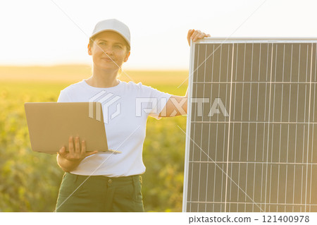 Woman farmer wearing white cap and t-shirt with laptop stands next to solar panel Woman farmer wearing white cap and t-shirt with laptop stands next to solar panel 121400978