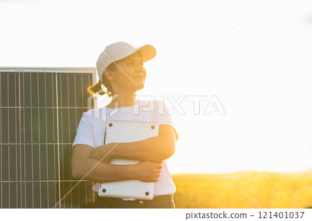 Woman farmer wearing white cap and t-shirt with laptop stands next to solar panel. 121401037