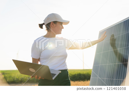 Woman farmer wearing white cap and t-shirt with laptop stands next to solar panel. Woman farmer wearing white cap and t-shirt with laptop stands next to solar panel. 121401038