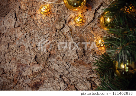 A wooden brown Christmas background with a spruce branch, garland, yellow and gold balls. Empty space for text. Happy New Year. The background is made of spruce bark. 121402953