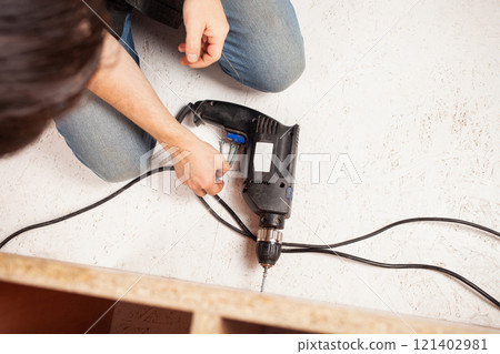 The man is holding screws in his hand, the drill is lying on the white floor. The man is holding screws in his hand, the drill is lying on the white floor. 121402981