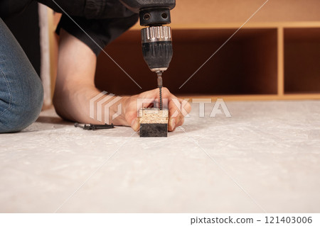 A man drills a hole in the wood against the background of an inverted closet on the floor. 121403006
