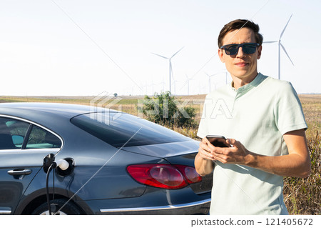 Man wearing sunglasses stands next to charging electric car and holds smartphone. Wind turbines in the background. Man wearing sunglasses stands next to charging electric car and holds smartphone. Wind turbines in the background. 121405672